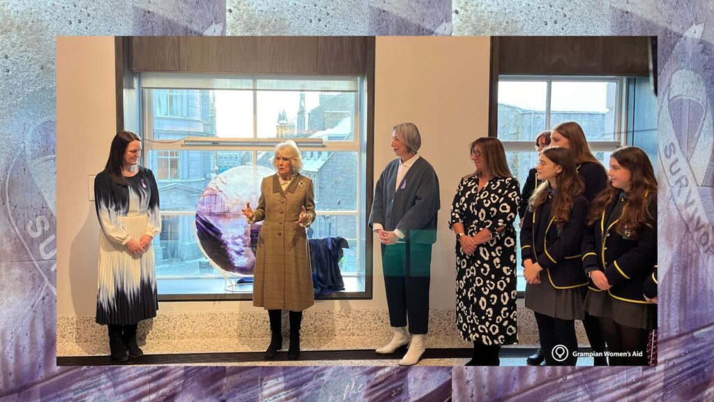 Queen Camilla talks to a group of assembled people. Behind her is a transparent and coloured circular artwork, with its cloth covering placed beside it
