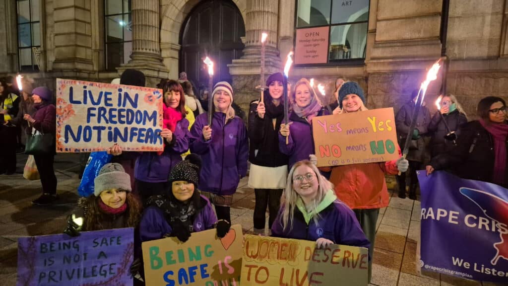A group of women hold protest placards and fire torches in the streets at night. The placards have messages such as "live in freedom not in fear" and "yes means yes, no means no"